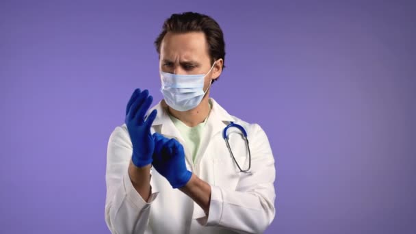 Mladý lékař si obléká latexové rukavice a připravuje se na pracovní operaci. Violet studio pozadí. Pohlední muži v ochranné masce obličeje a bílém plášti. — Stock video