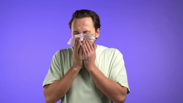ハンサムな男は組織にくしゃみをする。バイオレットスタジオの背景に孤立した男。彼は病気で、風邪やアレルギー反応があります。コロナウイルス流行2021病気の概念 — ストック動画