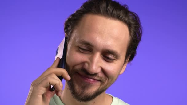 El joven habla con una sonrisa por teléfono. Un tipo sosteniendo y usando un teléfono inteligente. Estudio violeta fondo. — Vídeos de Stock