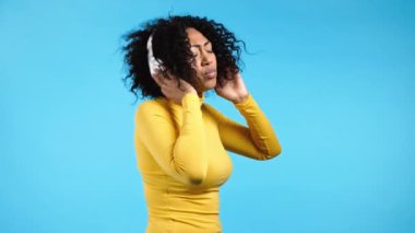 Mavi stüdyo duvarında izole edilmiş kablosuz kulaklıklarla dans eden Afrikalı kadın. Sarı elbiseli tatlı bayan, şarkı söylüyor. Müzik, radyo, mutluluk, özgürlük, gençlik kavramı.