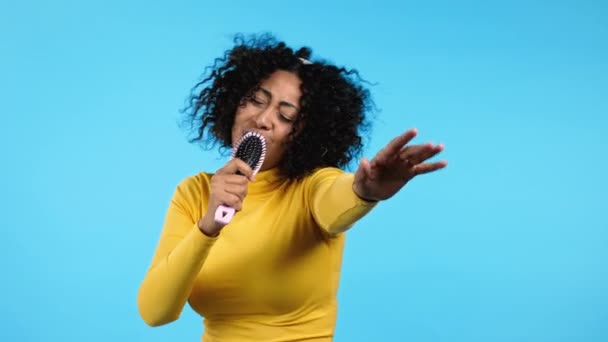 Африканська жінка співає і танцює з щіткою для волосся або гребінцем замість мікрофона біля блакитної стіни. Дама в навушниках розважається, слухає музику, мріє бути знаменитістю. — стокове відео