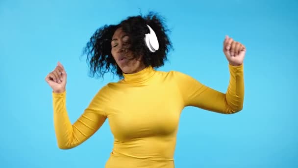 Vegyes rasszista nő táncol vezeték nélküli fejhallgatóval kék stúdió háttérben. Aranyos női portré. Zene, rádió, boldogság, szabadság, ifjúságpolitika. — Stock videók