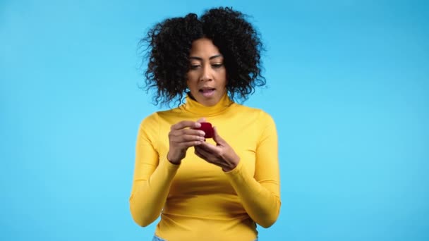 Attractive african woman holding small jewelry box with proposal diamond ring on blue wall background. Lady screaming, jumping , she is happy to get present, proposition for marriage — Stock Video