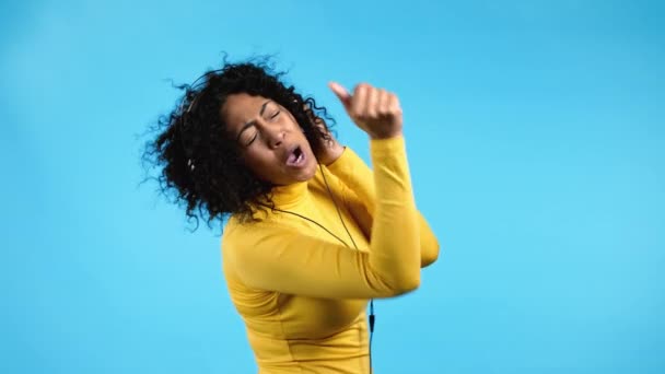 Mujer afroamericana bailando con auriculares sobre fondo azul. La chica se droga por la pista de música. Radio, felicidad, libertad, concepto de juventud. — Vídeo de stock