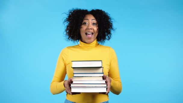 Estudiante afroamericano en ropa amarilla sostiene pila de libros universitarios de la biblioteca sobre fondo azul en el estudio. Mujer sonríe, ella está feliz de graduarse. — Vídeo de stock