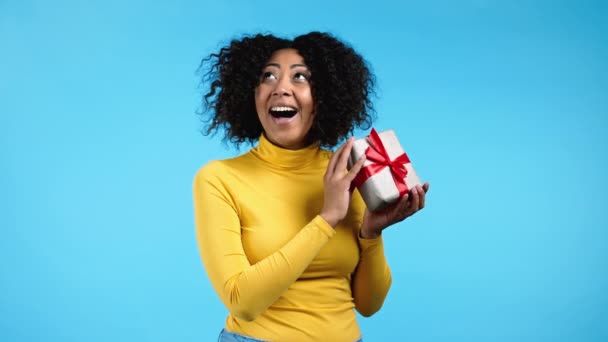 Mujer africana emocionada recibió caja de regalo con lazo. Ella está feliz y halagada por la atención. Chica sonriendo con presente sobre fondo azul. Retrato de estudio — Vídeo de stock