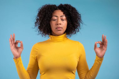 Mavi elbiseli sakin Afrikalı kadın dinleniyor, meditasyon yapıyor. Sakinleşiyor, derin derin derin nefes alıyor. Mavi stüdyo arka planında. Yoga konsepti.