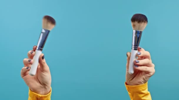 Vrouwelijke handen houden gezicht make-up borstels op geïsoleerde blauwe achtergrond. — Stockvideo