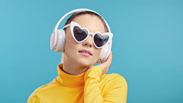 青いスタジオの背景に隔離されたワイヤレスヘッドフォンで踊るかなりの女性。ハート型のサングラスをかけた可愛い女の子。音楽、ラジオのコンセプト. — ストック動画