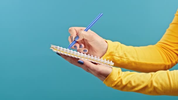 Manos de mujer tomando notas en el planificador, señora sosteniendo pluma. Ella escribe planes futuros y lista de tareas pendientes en un cuaderno para la semana o el mes. Mantener diario personal en el fondo del estudio azul. — Vídeos de Stock