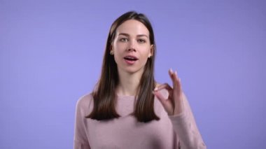 Genç bir kadın OK 'u gösteriyor, Violet stüdyosunun arka planını imzalamak gibi. Olumlu genç kız kameraya gülümsüyor. Kazandım. Başarı. Vücut dili.