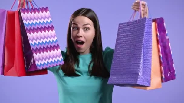 Excited woman with colorful paper bags after shopping on violet studio background. Concept of seasonal sale, purchases, spending money on gifts — Stock Video