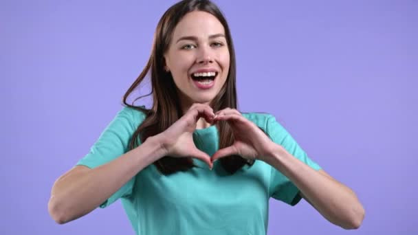 Bonita mujer haciendo señal de forma en el corazón cerca de su pecho. Fondo violeta. Salud de las mujeres, voluntariado, ayuda para la donación y concepto de amor. — Vídeos de Stock
