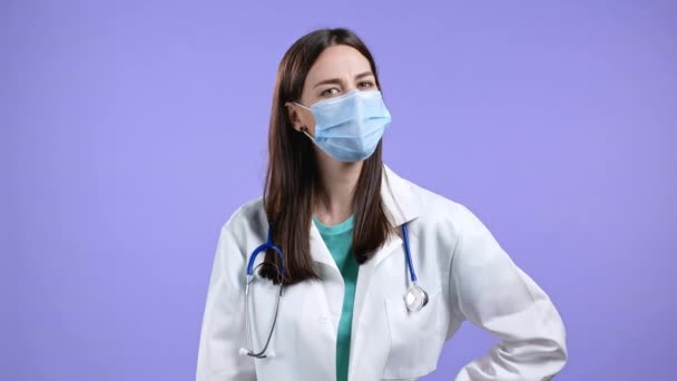 Portret van verpleegster in beschermende medische masker en jas met ja teken, knikt haar hoofd goedkeurend. Positieve dokter vrouw, lichaamstaal concept. Violette studio achtergrond. — Stockvideo