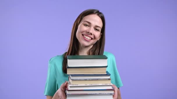 Student drží hromadu univerzitních knih z knihovny na fialovém pozadí ve studiu. Žena se usmívá, je šťastná, že odmaturovala. — Stock video