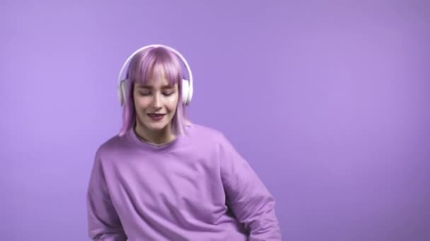 Красивая девушка с окрашенными фиолетовыми волосами танцует с беспроводными наушниками на фиолетовом фоне студии. Активный портрет женщины-хипстера. Музыка, радио, счастье, свобода, концепция молодости. — стоковое видео