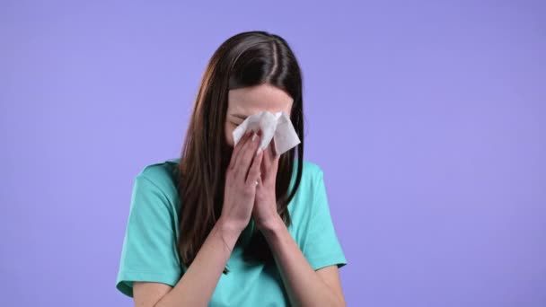 Mujer joven estornuda en el tejido. Chica aislada sobre fondo de estudio violeta. La señora está enferma, tiene un resfriado o una reacción alérgica. Coronavirus, epidemia 2021, concepto de enfermedad — Vídeos de Stock