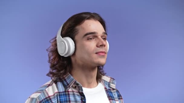 Encantador joven con el pelo largo escuchando música con auriculares inalámbricos, chico divirtiéndose, sonriendo en el estudio sobre fondo violeta. Danza, concepto de radio. — Vídeos de Stock