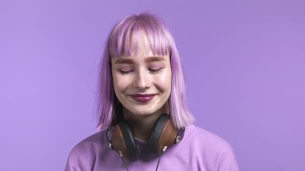 Портрет жінки-гіпстера з пофарбованим фіолетовим волоссям на фіолетовому студійному фоні. Позитивна молода дівчина вечірки з навушниками на шиї дивиться на камеру. Дивовижний макіяж і чарівна посмішка . — стокове відео