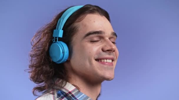 Όμορφος νεαρός άνδρας που ακούει μουσική με ασύρματα ακουστικά, τύπος με σγουρά μακριά μαλλιά που διασκεδάζει, χαμογελώντας στο στούντιο σε βιολετί φόντο. Χορός, ραδιοφωνική αντίληψη. — Αρχείο Βίντεο