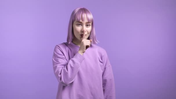 Usmívající se žena s obarvenými vlasy, držící si prst na rtech na fialovém pozadí. Gesto shhh, tajemství, ticho. Zavřít. — Stock video