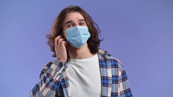 Bărbat mulțumit cu păr lung îndepărtat masca de protecție facială ca simbol al pandemiei, capăt epidemic. Violet studio background — Videoclip de stoc