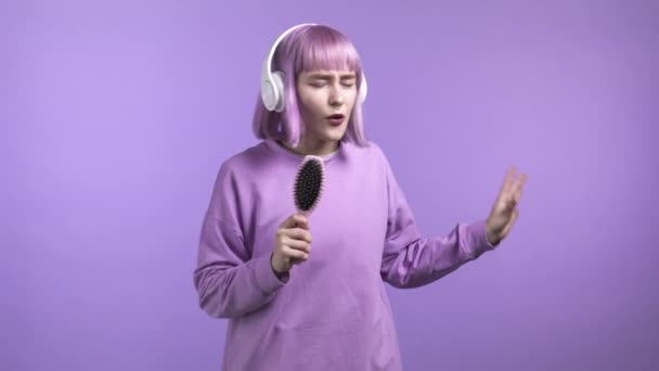 Hezká dívka s obarvenými fialovými vlasy zpívající a tančící štětcem nebo hřebenem místo mikrofonu na fialové stěně. Žena ve sluchátkách se baví, poslouchá hudbu, sní o tom být celebritou. — Stock video
