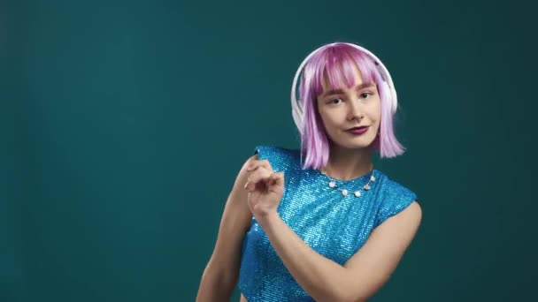 青いスタジオの背景にヘッドフォンで踊る魅力的な現代女性。かわいい紫染めの髪の女の子の肖像画。音楽、ラジオ、パーティーのコンセプト. — ストック動画