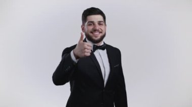 Smokinli sakallı yakışıklı adam başparmağını kaldırıyor. Beyaz arka plandaki mutlu adam. Kazandım. Başarı. Pozitif erkek model kameraya gülümser. Vücut dili. 