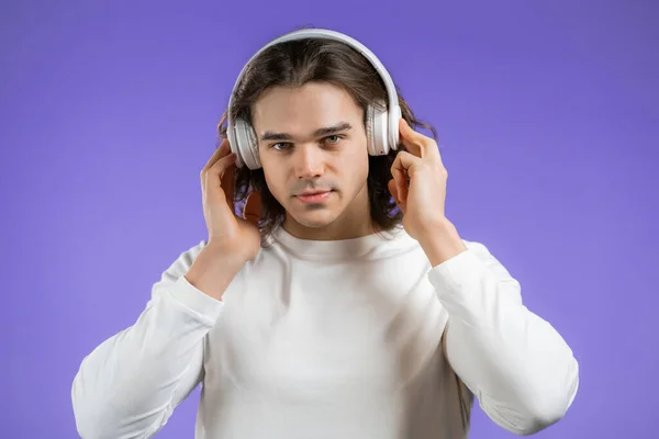 Charmig ung man med lång frisyr lyssnar på musik med trådlösa hörlurar, killen har roligt, ler i studio på violett bakgrund. Dans, radiokoncept. — Stockfoto
