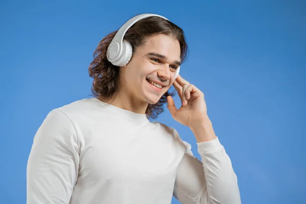 Charmig ung man med lång frisyr lyssnar på musik med trådlösa hörlurar, killen har roligt, ler i studio på blå bakgrund. Dans, radiokoncept. — Stockfoto