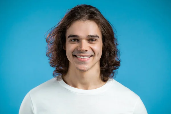 Retrato de homem europeu alegre olhando para câmera. Jovem bonito tipo cara com longo penteado sorrindo em estúdio no fundo azul. — Fotografia de Stock