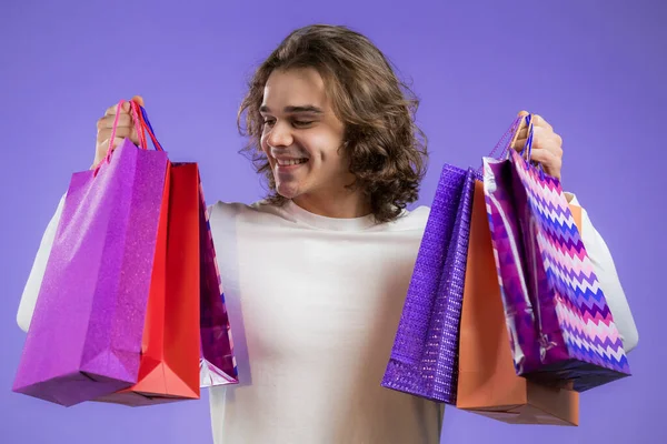 Schöner Mann mit langer Frisur hält Einkaufstüten auf violettem Studiohintergrund. Kerl kaufte Geschenke mit Rabatten. — Stockfoto