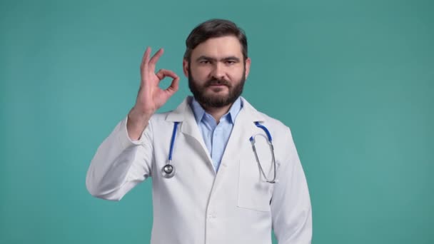 Doctorul arată bine, bine ca un semn pe fundal albastru. Un tânăr cu haină medicală zâmbește camerei. Câştigător. Succes. Limbajul corpului . — Videoclip de stoc