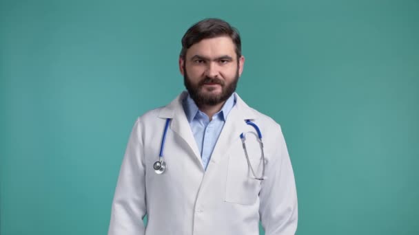 Hombre guapo doctor en bata blanca profesional sobre fondo de estudio azul. Doctor serio con barba mirando a la cámara. — Vídeo de stock
