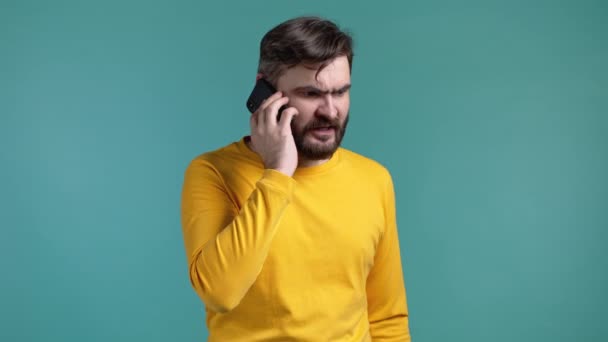 Angry bearded man in yellow t-shirt screaming down his mobile phone. Stressed and depressed guy on blue background. — Stock Video