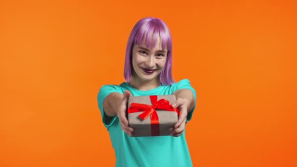 Mujer con pelo violeta da caja de regalo con las manos a la cámara en fondo de estudio naranja. Chica sonriendo, ella es feliz con el presente. — Vídeo de stock