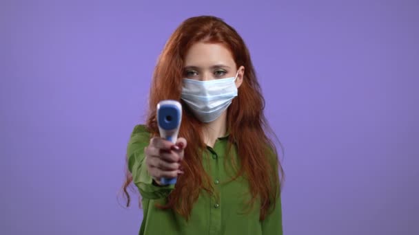 Mujer europea en máscara protectora dirige termómetro electrónico sin contacto en la lente de la cámara y mide la temperatura. — Vídeo de stock