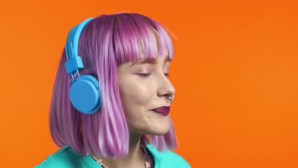 Attraktiv modern kvinna dansar med hörlurar på orange studio bakgrund. Söta tjejer med violett färgat hår porträtt. Musik, radio, lycka koncept. — Stockvideo