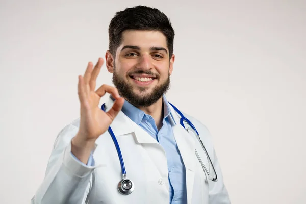 Doktor toont ok, oke als teken over witte achtergrond. Positieve jongeman in medische jas glimlacht naar de camera. Winnaar. Succes. Lichaamstaal. — Stockfoto