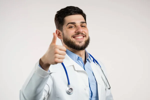 Arzt zeigt Daumen-hoch-Zeichen über weißem Hintergrund. Positiver junger Mann im Arztkittel lächelt in die Kamera. Sieger. Mit Erfolg. Körpersprache. — Stockfoto