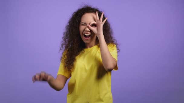 Hübsche Frau zeigt ok, okay Zeichen auf violettem Studiohintergrund. Positives junges Mädchen mit lockigem Haar lächelt in die Kamera, hat Spaß, macht Brille aus Fingern — Stockvideo