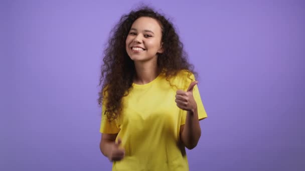 Vacker kvinna visar tummen upp som skylt över violett studio bakgrund. Positiv ung flicka med lockigt hår ler mot kameran. Vinnare. Framgång. Kroppsspråk. — Stockvideo