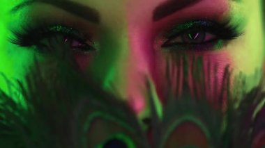 Tavus kuşu tüylü bir kadın yüzü. Güzel makyajlı, ışıl ışıl göz farları olan bir kadın. Yeşil gözlü ve takma kirpikli bir kız. Güzellik, kozmetik, sanat, kadınsı konsept.