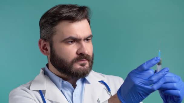 Доктор в перчатках щёлкает шприцем перед введением препарата, чтобы собрать и выпустить воздух из шприца. Бородатый мужчина на фоне синей студии. Здравоохранение, вакцинация, концепция медицины. — стоковое видео