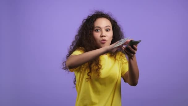 Curly haired woman with happy face scatters money. Lady is overspend USD currency. Girl is flush with dollars on violet studio background. — Stock Video