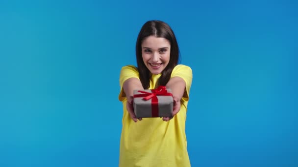 Femme aux cheveux longs donne boîte cadeau à la main à la caméra sur fond de studio bleu. Fille souriante, elle est heureuse avec le cadeau. — Video