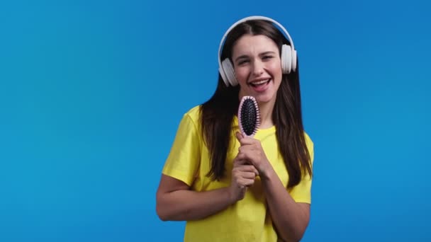 Mujer europea cantando y bailando con cepillo de pelo o peine en lugar de micrófono en fondo de estudio azul. Dama en auriculares divirtiéndose, escuchando música, sueña con ser celebridad. — Vídeos de Stock