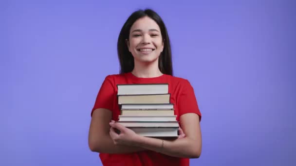 Студентка держит стопку университетских книг из библиотеки на фиолетовом фоне в студии. Женщина улыбается, она счастлива выпуститься. — стоковое видео