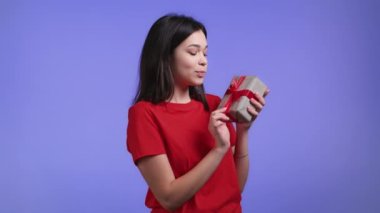 Heyecanlı kadın fiyonklu hediye kutusu aldı. O mutlu ve ilgiyle pohpohlanıyor. Menekşe arkaplanda gülümseyen bir kız. Stüdyo resmi